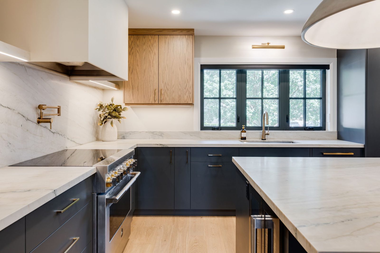 Kitchen run with white oak upper cabinets, navy base cabinets, a stone slab backsplash, a stainless pro range and brass hardware.
