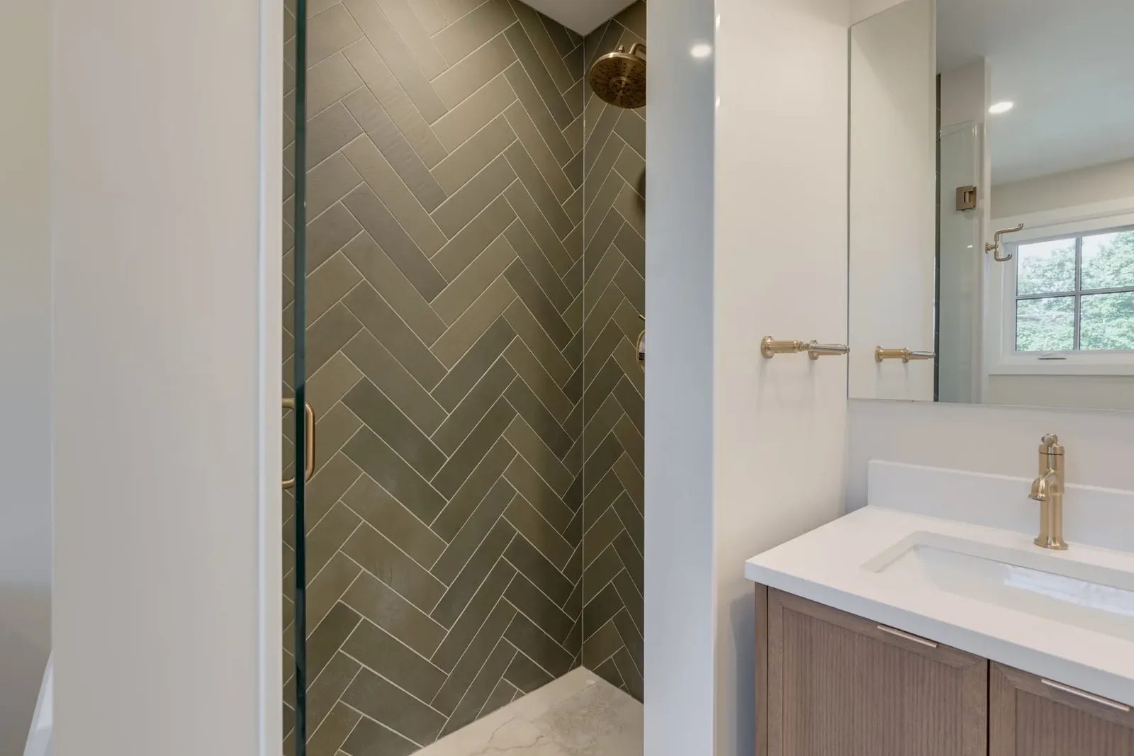 Curbless walk-in shower clad in dark green herringbone tile behind a frameless glass panel, with the walnut vanity beyond.