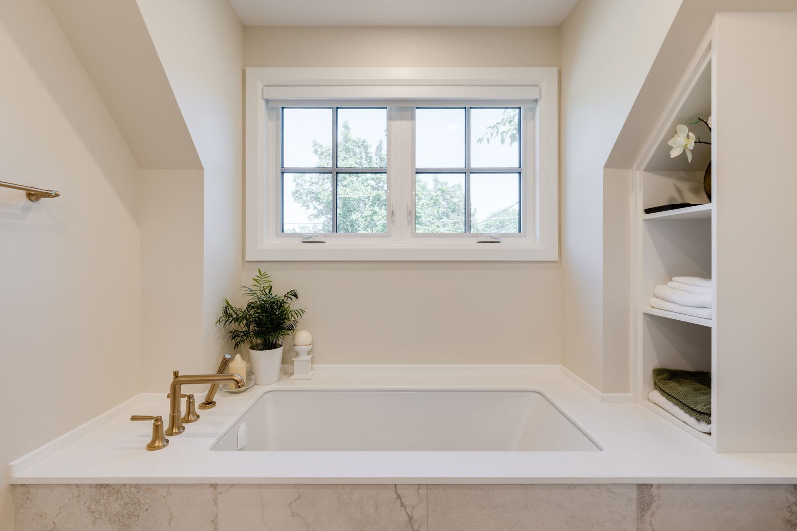 Stone tub deck built into the alcove with a brass deck-mounted filler, beside recessed white linen shelving.