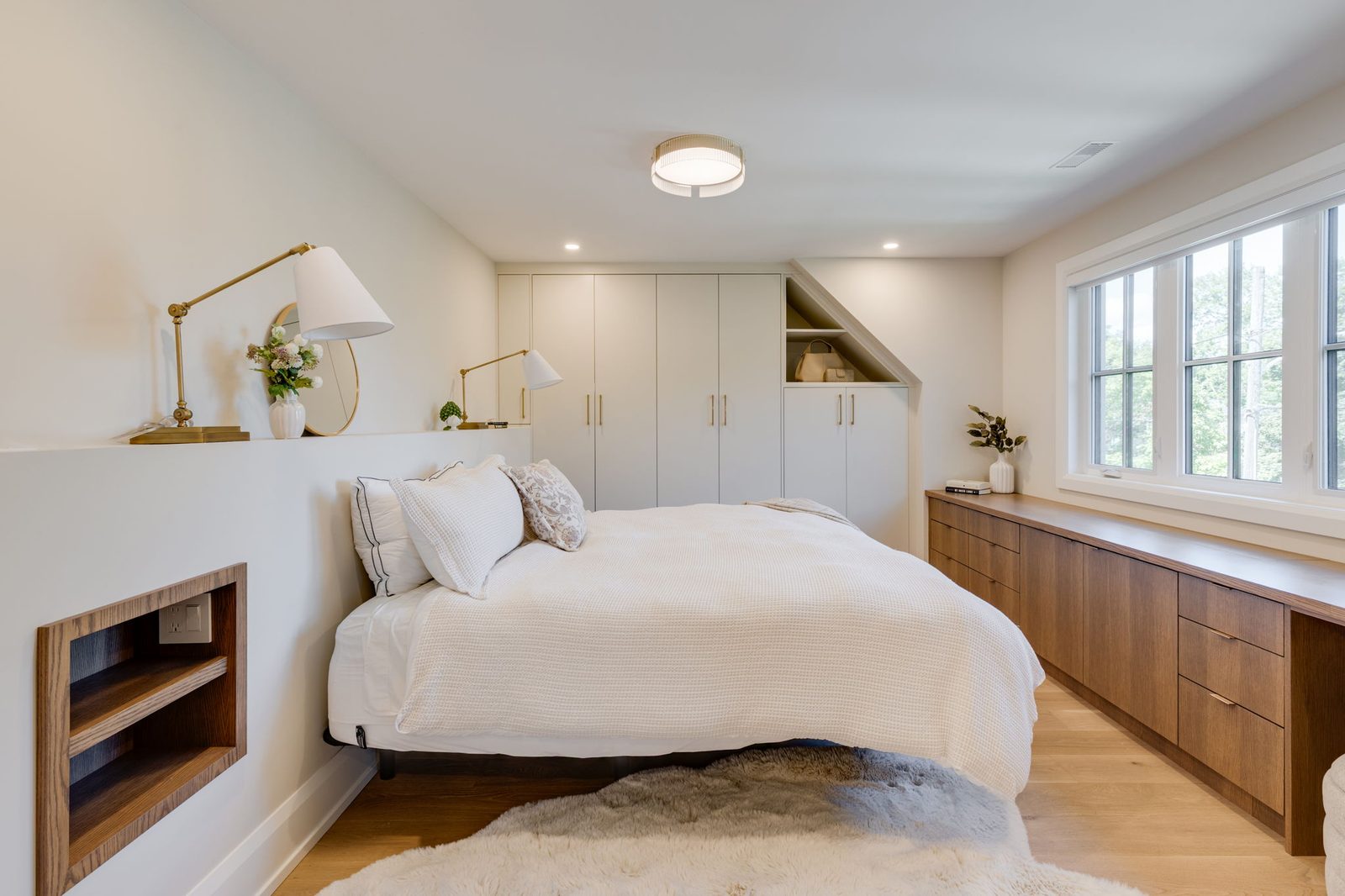 Bedroom with a wall of white built-in wardrobes, a walnut dresser run under the window and a built-in headboard niche.